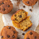 Five chocolate chip pumpkin muffins surrounding one pumpkin muffin that is broken in half with the inside facing up. The muffins are on a piece of parchment paper on wood.