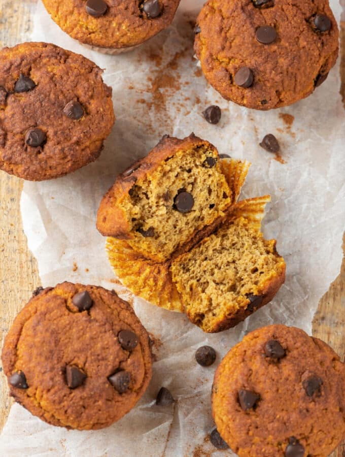 Five chocolate chip pumpkin muffins surrounding one pumpkin muffin that is broken in half with the inside facing up. The muffins are on a piece of parchment paper on wood.