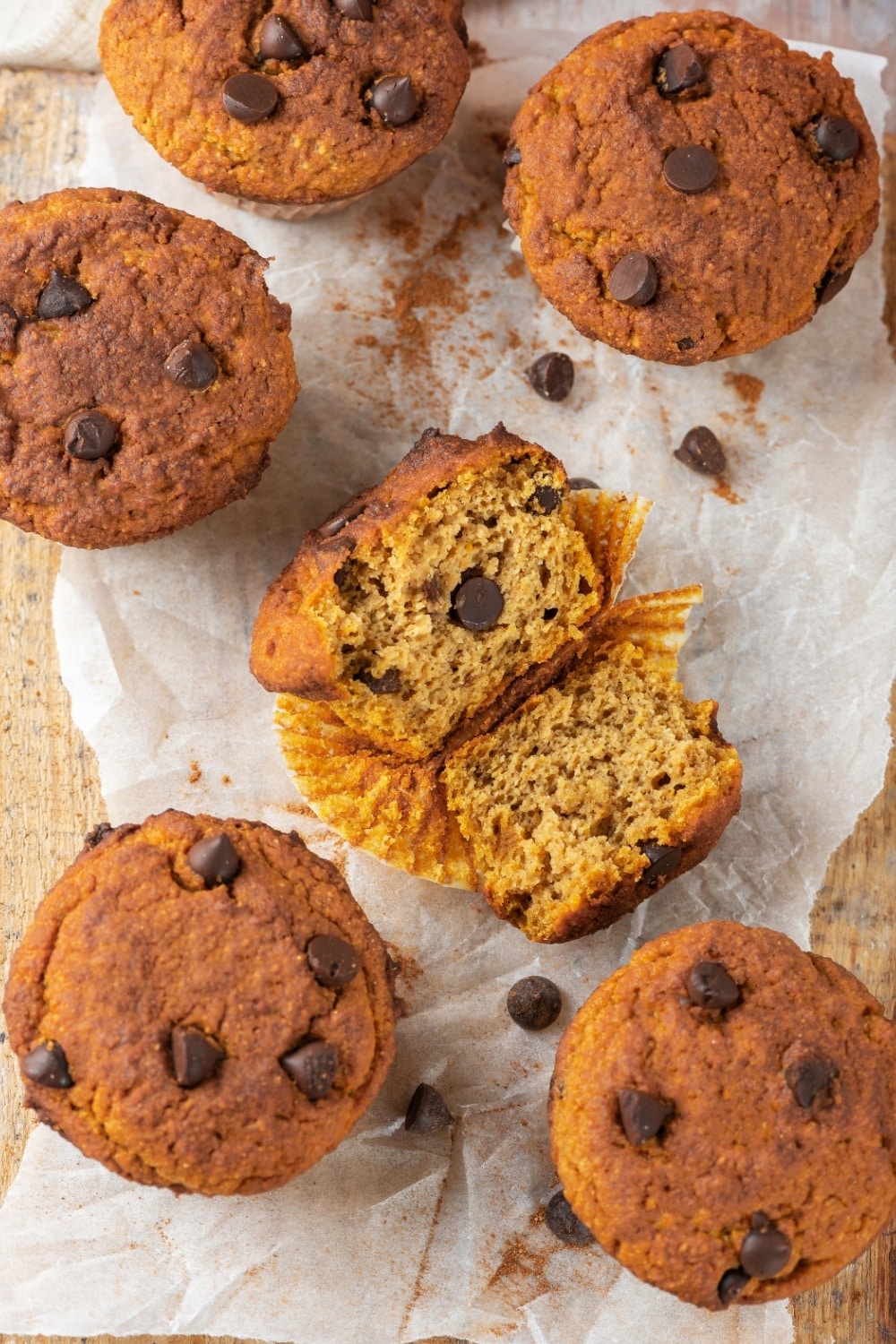 Five chocolate chip pumpkin muffins surrounding one pumpkin muffin that is broken in half with the inside facing up. The muffins are on a piece of parchment paper on wood.