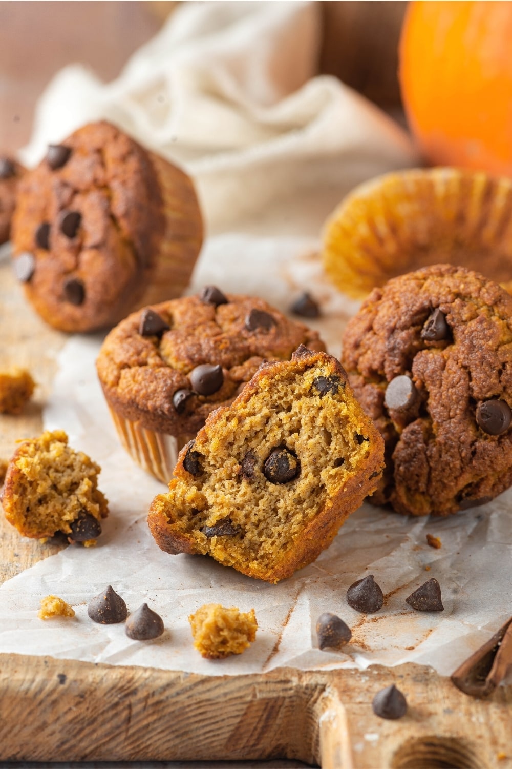 A couple of chocolate chip pumpkin muffins and a piece of parchment paper on a morning counter. There are some chocolate chips surrounding the muffins and one of the muffins in the front is cut in half with inside showing.