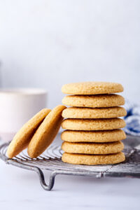 Seven shortbread cookies stacked on top of one another on a wire rack on the white counter. There are two shortbread cookies leaning against the left of the stack. Behind that is a white coffee cup.