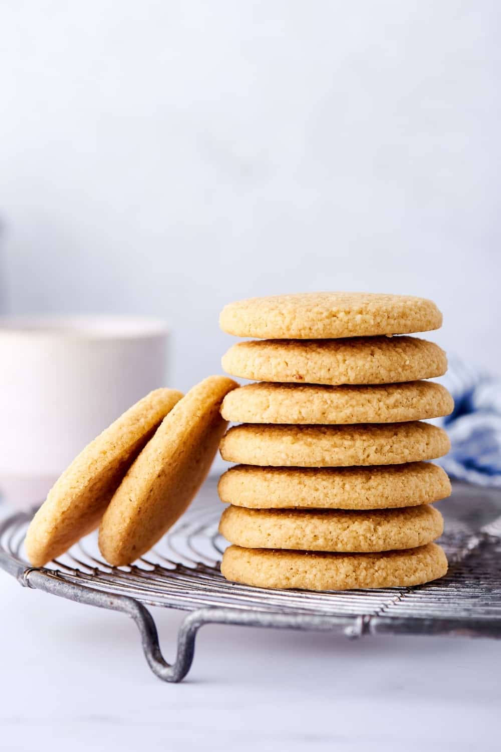 Seven shortbread cookies stacked on top of one another on a wire rack on the white counter. There are two shortbread cookies leaning against the left of the stack. Behind that is a white coffee cup.