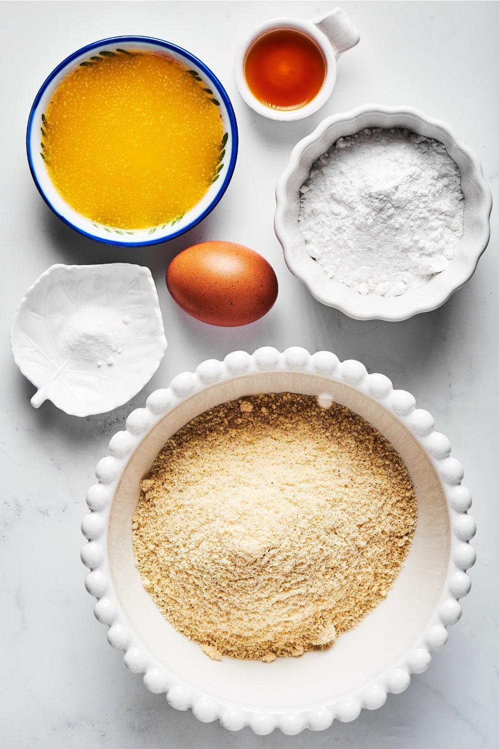 A white bowl of almond flour, a small leaf dish with salt Nick, an egg, bowl of powdered erythritol, aa bowl of melted butter. and a small bowl of vanilla extract all on a white counter.