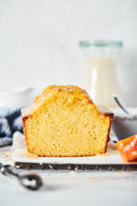 A poundcake loaf on a piece of white parchment paper on a black wire rack on top of the white counter. There's a slice out of the front of the poundcake and a tiny piece of that slice is at the edge of the parchment paper. Behind the loaf is a glass jar of milk