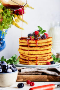 A cream cheese pancakes stacked on top of one another on a white plate that is on a gray tablecloth and a wooden cutting board and a white counter. A glass pitcher is drizzling syrup on top of the pancakes and there are a few blueberries and raspberries on the top pancake.