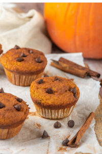 Three pumpkin muffins and a piece of parchment paper with some chocolate chips and cinnamon sticks on it. Behind a piece of parchment paper as part of a pumpkin.