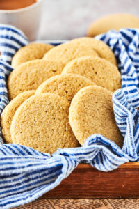 A wooden basket with a blue and white striped tablecloth in it with a bunch of shortbread cookies overlapping one another.