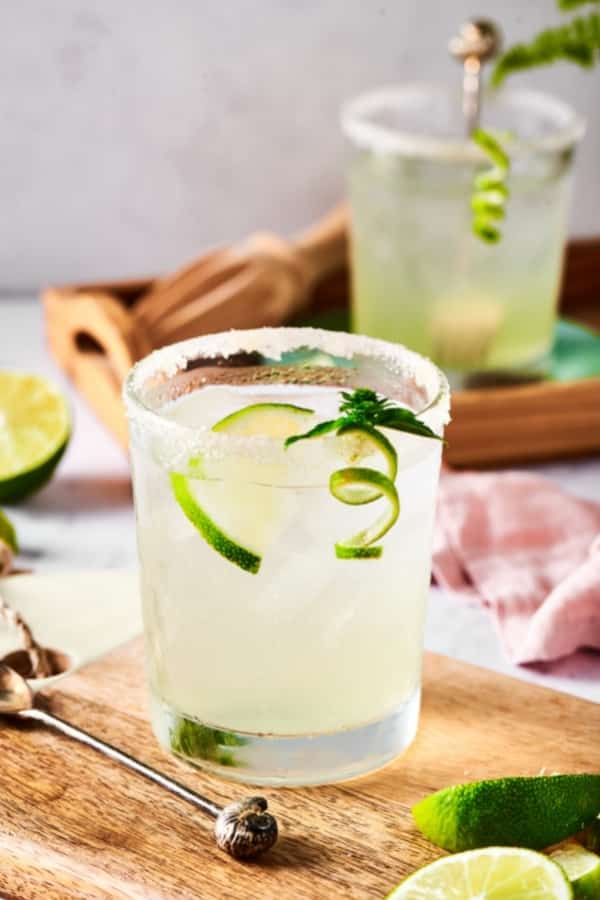 A wooden board with a glass filled with margarita on it. There is a lime wedge submerged in the margarita and a few lime wedges in front of the glass of margarita. Behind it is a wooden serving tray with another glass of margarita on it.