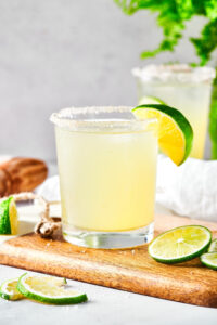 A wooden board on a white counter with a glass of margarita on it there's a lime wedge on the rim of the glass into the right of a glass or a few lime slices. Behind it is another glass of margarita.