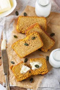 Four slices of pumpkin bread scattered on a piece of brown parchment paper and a gray tablecloth. The front slice is cut in half and has a slab of butter on top and there is a knife next to it with butter on it. To the right of that is a glass jar of milk and behind the last slice of bread is another glass jar of milk.