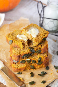 A stack of four slices of pumpkin bread and a piece of brown parchment paper on a gray tablecloth. The top slice is cut in half and there is a slab of butter on top of it..