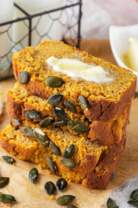 Four slices of pumpkin bread stacked on top of one another on a piece of parchment paper. The top slice has a slab of butter on it and pumpkin seeds in front of the bread.