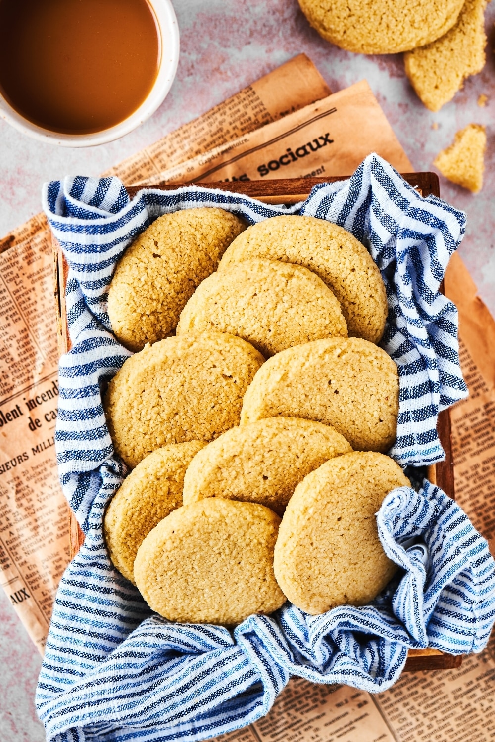 Brown newspaper on a white counter with a basket filled with a bunch of shortbread cookies overlapping one another in it. The shortbread cookies are on a white and blue striped tablecloth and behind it is part of a cup of coffee and parts of other shortbread cookies.