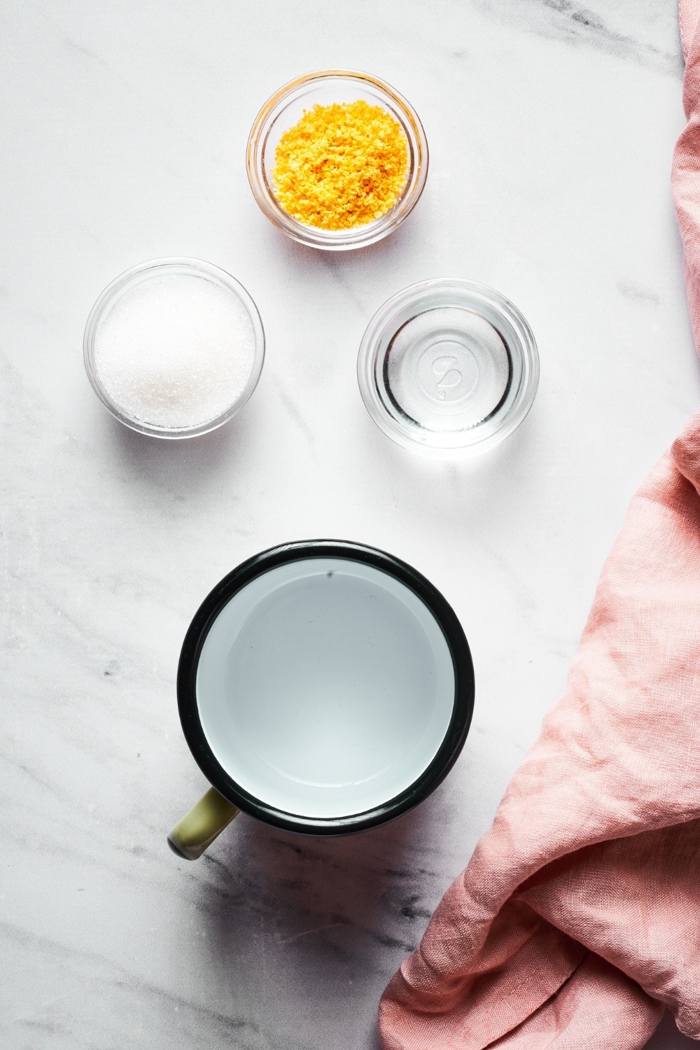 An empty cup, a small bowl of erythritol, a small bowl of water, and a small bowl of orange sauce all on a white counter.