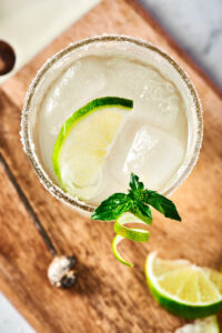 Wooden board with a glass filled with margarita on it. There's a lime wedge on top of the ice cubes in the glass and there is one lime wedge in front of the glass on the wooden board.