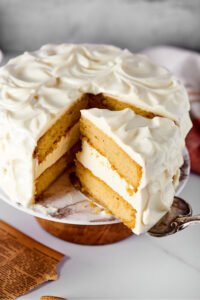 A vanilla cake with vanilla frosting on a cake stand. A cake spatula is pulling a slice of cake from it.