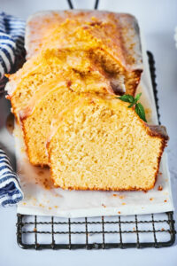 Pound cake loaf on a piece of parchment paper on top of a black wire rack on a white counter. There are three slices of poundcake staggered against one another leaning against the front of the loaf.