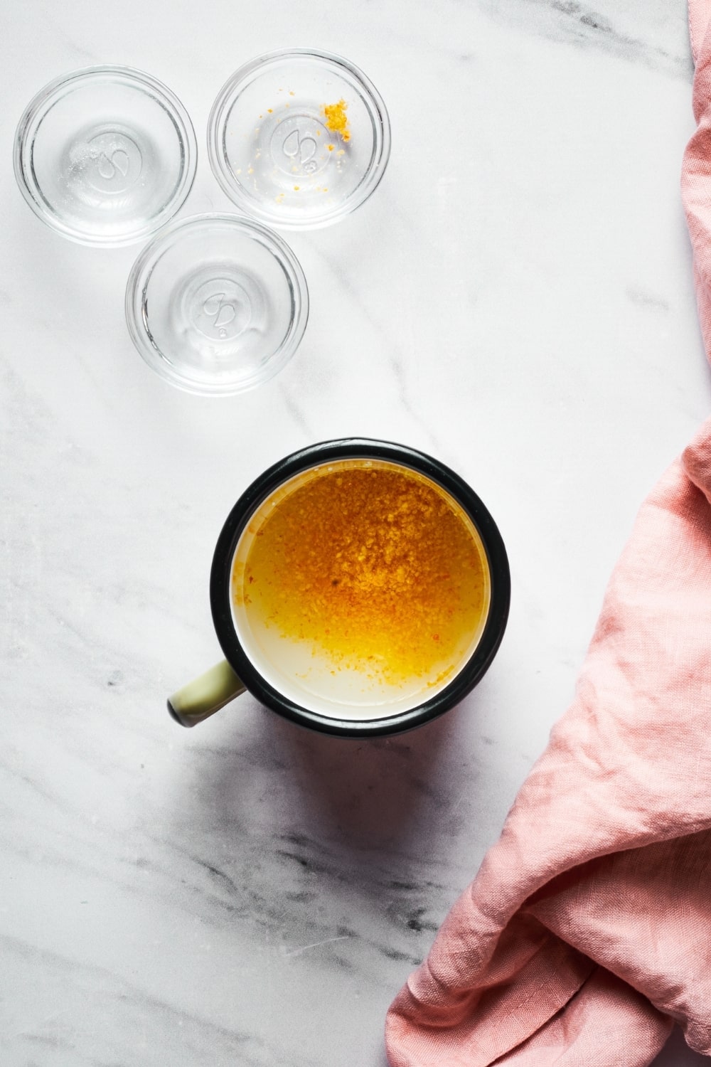 A cup filled with orange zest, water, and erythritol on a white counter. In front of it is three empty small glass bowls.