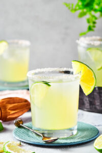 A glass of margarita on a circular blue coaster on a white counter. Behind it are parts of two other glasses of margarita.