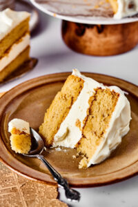 A slice of vanilla cake and a brown plate on a white counter. Hey spoons to the left of the piece of cake with part of the cake on it.