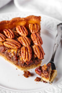 "A slice of pecan pie on a white plate. A piece of the front of the pie is on a fork directly in front of the pie on the plate.