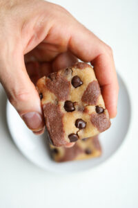 A hand holding an unbaked brookie over a white plate that has a brookie on it.