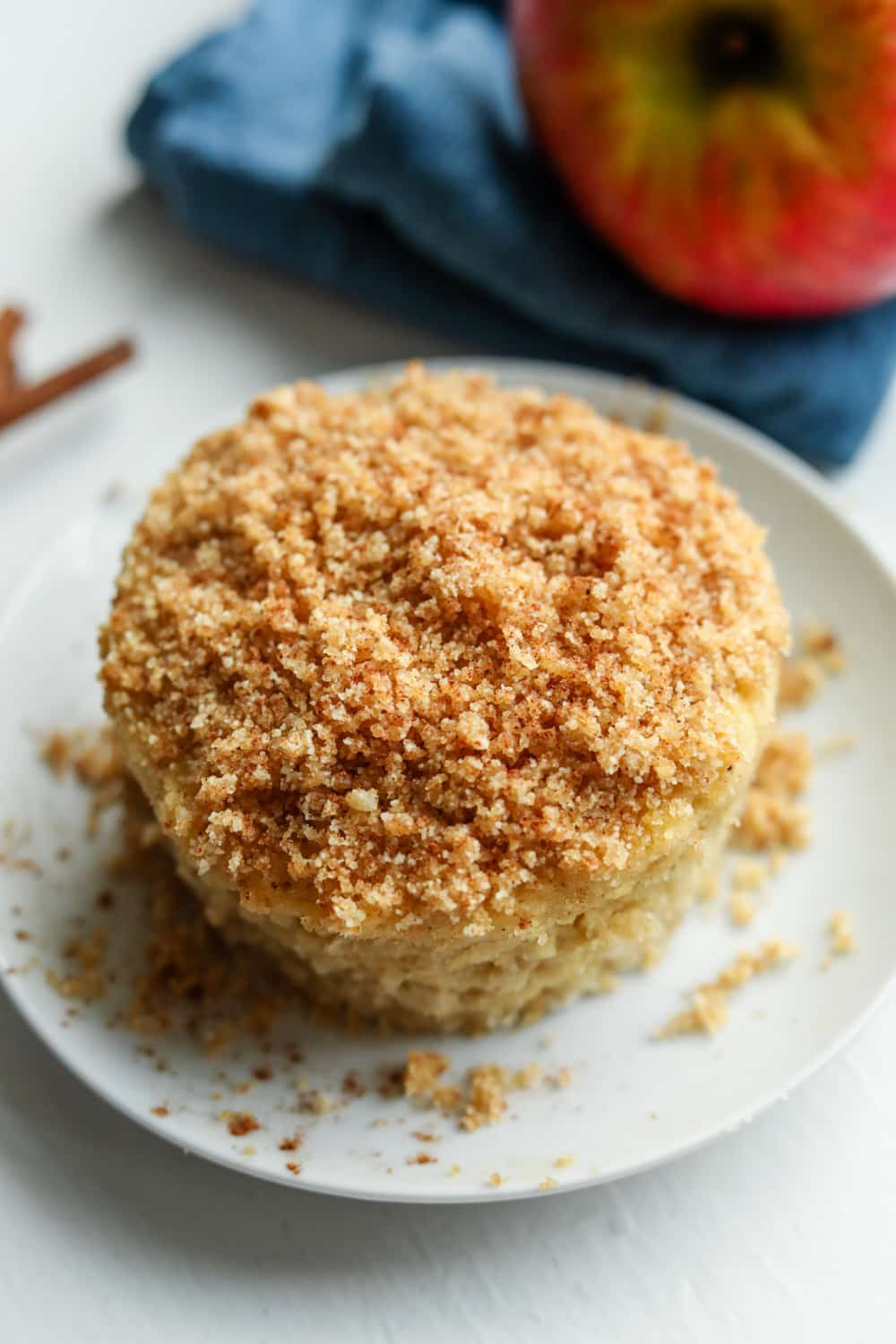 An apple crumble spice cake on a white plate. There is a blue napkin with an apple on top of it behind the cake.