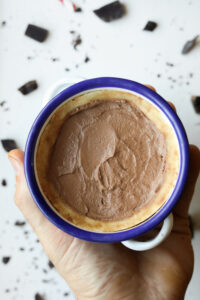 A chocolate pie inside of a bowl with a blue rim. There are pieces of chocolate on a white table underneath it.