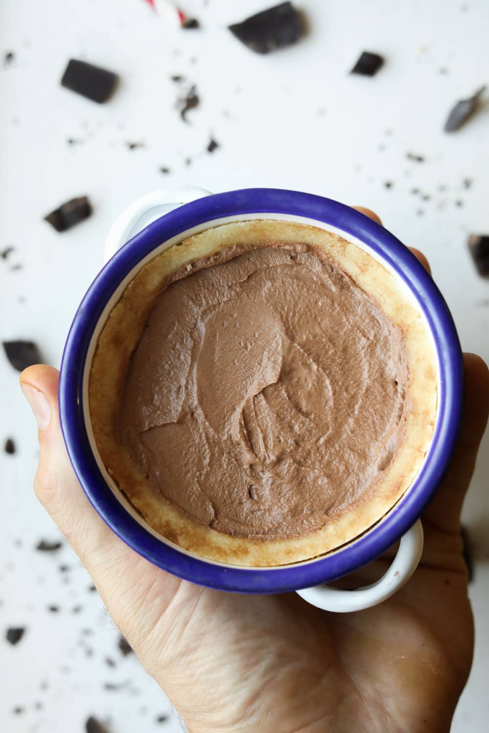 A chocolate pie inside of a bowl with a blue rim. There are pieces of chocolate on a white table underneath it.