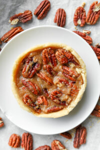 A mini pecan pie on top of a white dish on a grey counter surrounded by pecans.