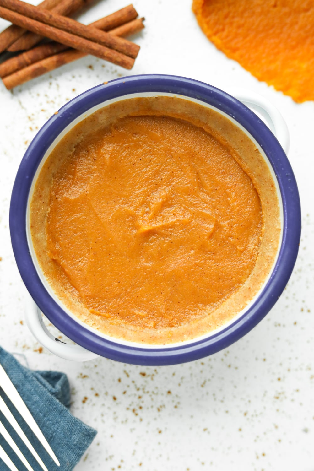 Pumpkin pie inside of a white bowl with the blue. There are cinnamon sticks, pumpkin purée, and a blue napkin next to the bowl.