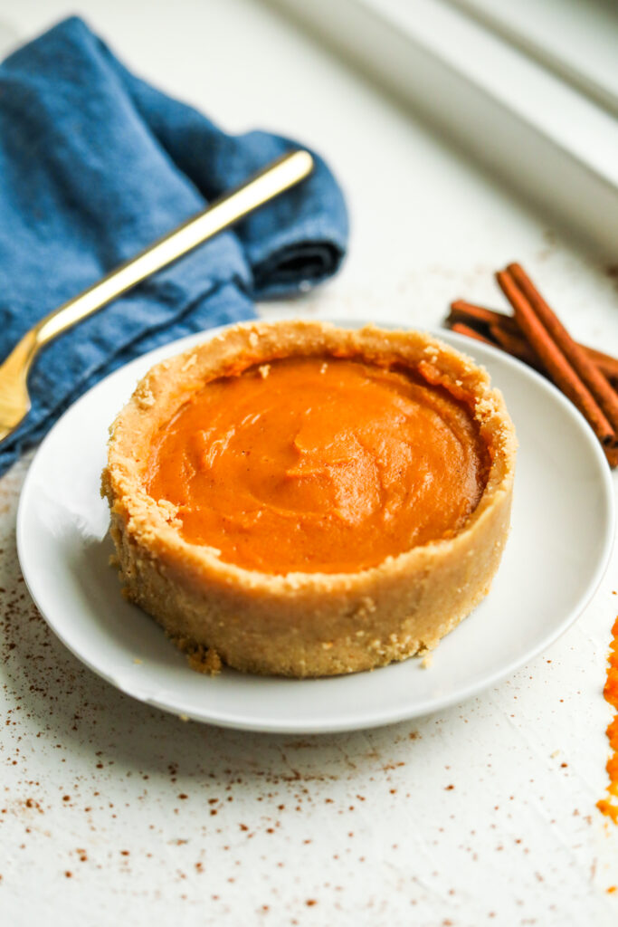 Pumpkin pie on a white plate. There are cinnamon sticks next to the plate along with a blue napkin and a gold fork.