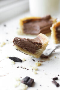 A fork holding a piece of chocolate pie. There is more of the pie behind it and a glass of milk behind the pie as well.