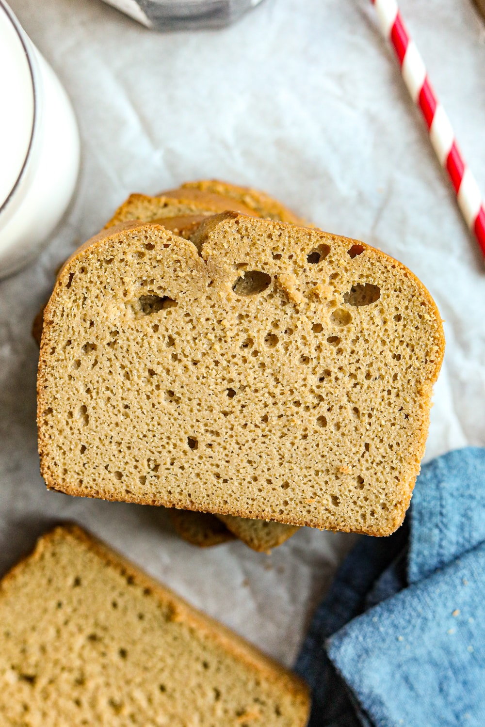 Slices of nut butter bread stacked on top of each other on a white sheet of parchment paper. There's a blue napkin, a red and white straw, and a glass of milk next to the bread.