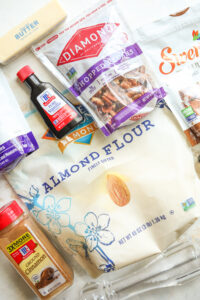 A bag of almond flour, a bag of chopped pecans, a bottle of vanilla extract, and a stick of butter on a white counter.