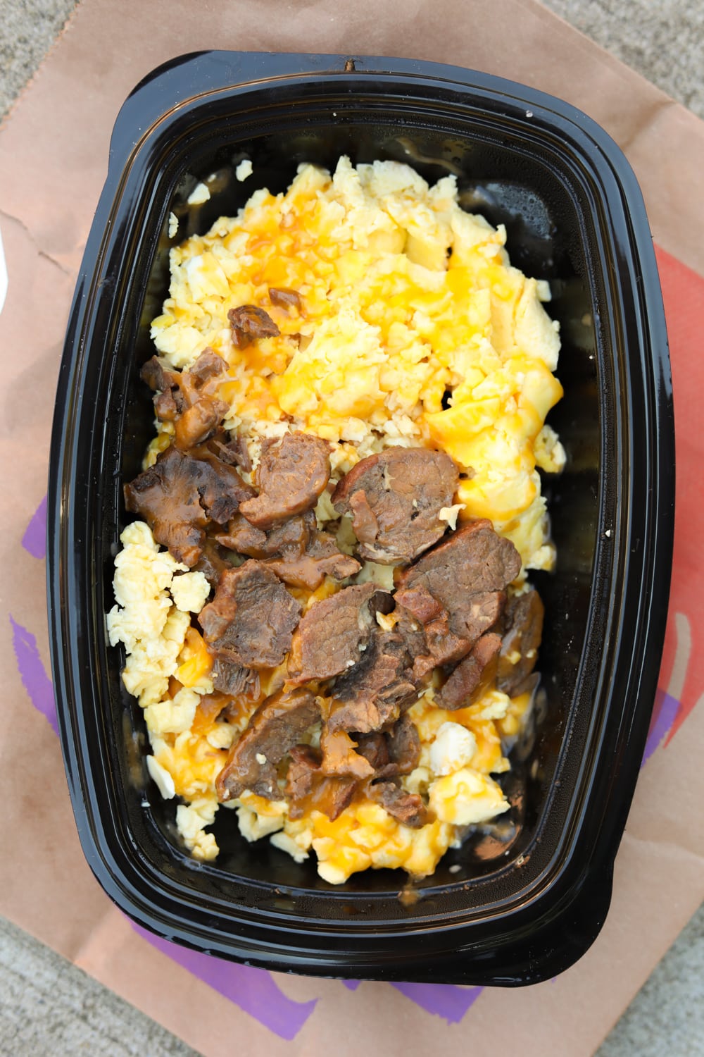 A black container with steak, egg, and cheese in it. The container is on a brown paper bag.