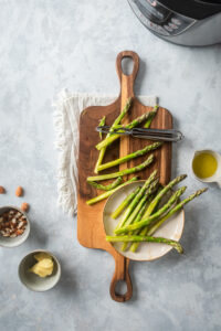 A white plate with asparagus on it on top of a wooden cutting board with some asparagus on it on a white tablecloth on a gray counter.