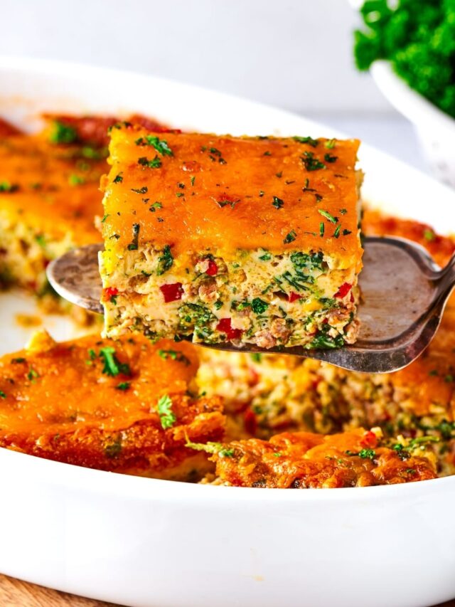 Hey square of breakfast casserole on top of a silver serving spatula hovering over a white casserole dish filled with breakfast casserole.