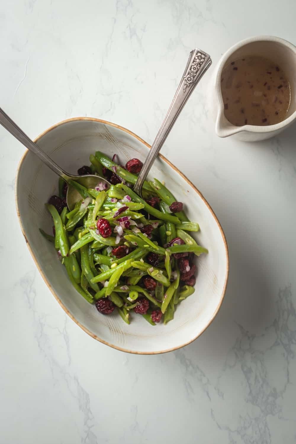 A white dish filled with green beans, red onion, and cranberries. Next to it is a small white pitcher filled with dressing.