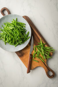 Green beans on a plate on a wooden cutting board on top of a white counter. There are a few more green beans next a knife on the wooden cutting board.