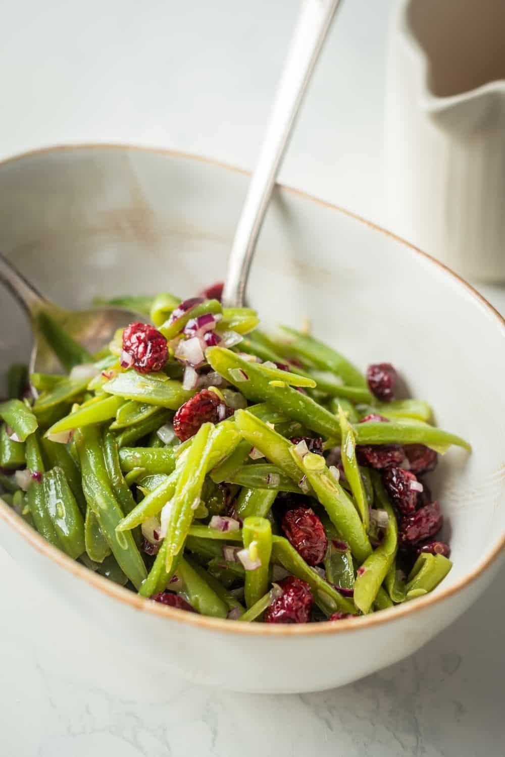 A white dish with green beans, red onion, and cranberries on top. There are two spoons in the green beans.