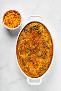 A breakfast casserole in a white casserole dish. At the back left of the dish is a small white bowl with shredded cheddar cheese in it.