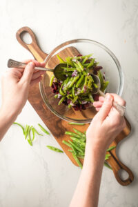 A glass bowl filled with green beans, red onion, and cranberries. There are two hands holding spoons in the bowl and the bowl is on a wooden cutting board.