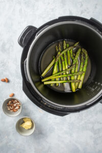 An instant pot filled with asparagus. Next to it is a small white bowl of almonds and a small white bowl of butter all on a gray counter.