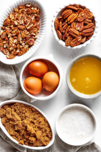 Ingredients for a sugar-free pecan pie filling on a white counter. There is a bowl of brown swerve, a bowl of full fat coconut milk, a bowl three eggs, a bowl of melted butter, a bowl with pecan halves, and a bowl of chopped pecans.