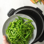 A hand holding a plate with green beans on it hovering over an instant pot.