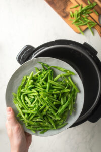 A hand holding a plate with green beans on it hovering over an instant pot.