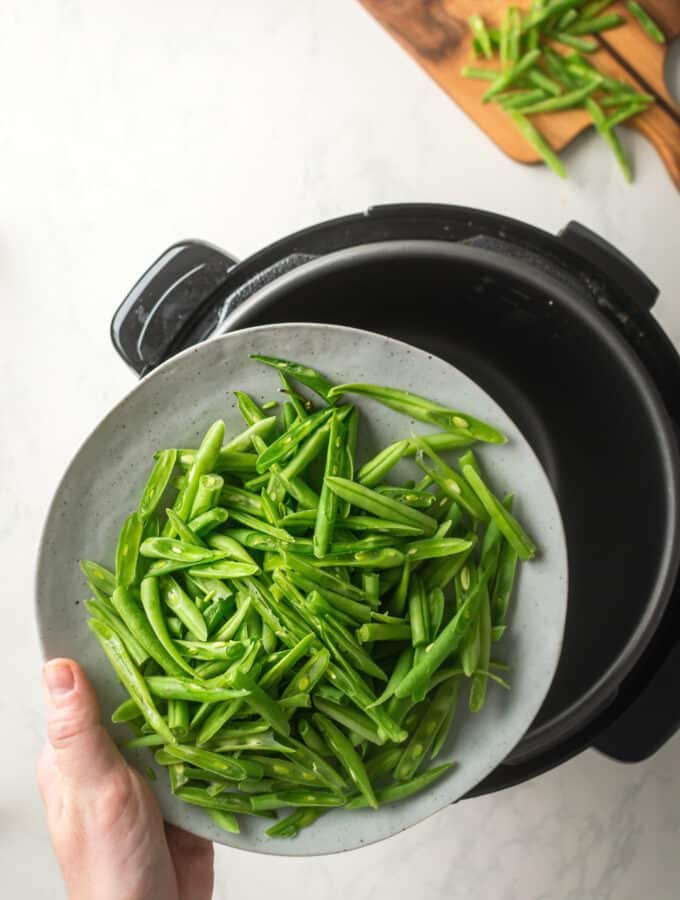 A hand holding a plate with green beans on it hovering over an instant pot.