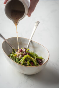 A white dish filled with green beans with red onion and cranberries on top. A hand is pouring dressing from a white pitcher onto the green beans.
