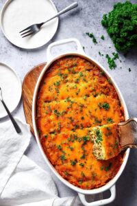 A white casserole dish with a breakfast casserole in it. There is a slice of the casserole being scooped out from the bottom right by serving spatula. The casserole dish is on a wooden cutting board and behind it is a white plate with a fork on it.